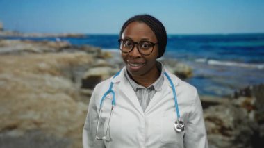 Plajda, steteskopuyla sakin deniz manzarasına bakan beyaz önlüklü bir kadın doktor rahatlamanın tadını çıkarıyor..