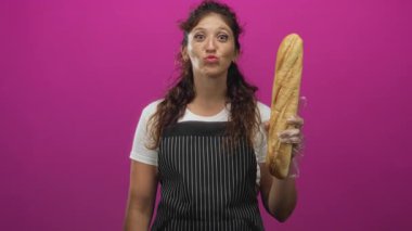 Dudaklarını büzen bir kadın eldivenli bir elle baget tutuyor. Stüdyonun canlı pembe arka planında çizgili önlük takıyor. Eğlenceli bir yemek pişirme..