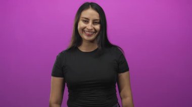 Genç bir kadın gülümsüyor ve mor stüdyoda el hareketi yapıyor. Siyah bir tişört ve görünür bir el ve kol giyiyor..