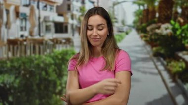 Pembe tişörtlü genç bir kadın güneşli bir sokakta hareketler yapıyor, canlı bir şehir ortamının ortasında ihtiyatlı bir ifade sergiliyor..