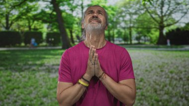 Elleri sımsıkı bağlı bir adam yeşil bir orman parkında dua ediyor, uzun gri saç ve sakal görünür; umut dua inanç.