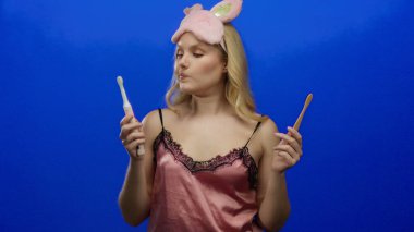 Gecelikli genç sarışın kadın mavi arka planda diş fırçası tutuyor neşeli ifadeleri ve uyku maskesiyle elektrik ve bambu seçenekleriyle çelişiyor.