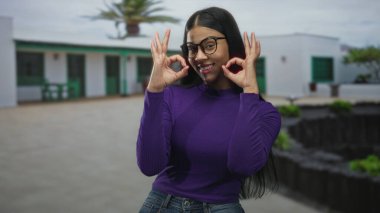 Kadın beyaz bina avlusunun önünde gülümserken iki eliyle tamam işareti yapıyor; teşvik desteği iyimserlik onayı.