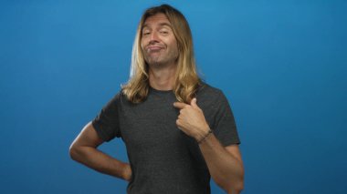 Sarışın, uzun saçlı, gri tişörtlü, bir eli kalçasında, mavi arka planlı, göğüs göğse gezen bir adam..