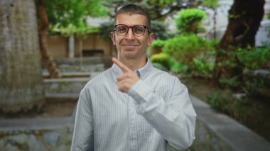 Parkta, yeşil ağaçlarla çevrili gözlüklü ve çizgili tişörtlü, merakla yukarıya bakan beyaz bir adam..