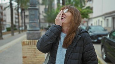 Şehir caddesinde gülen bir kadın araba ve ağaçlarla dolu, serin bir günde açık havada gözlük ve ceket takıyor..