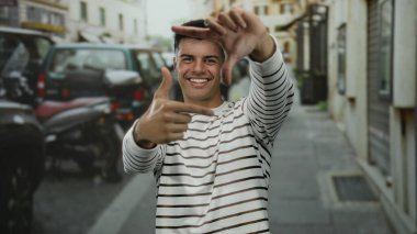 Çizgili tişörtlü genç adam gülümsüyor ve elleri ile şehir dışında canlı bir sokak arkaplanında çerçeveleniyor.