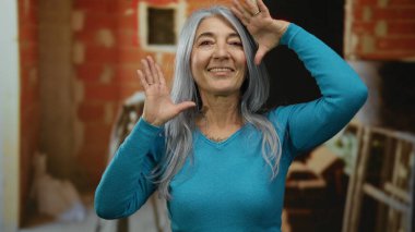 Gri saçlı, gülümseyen ve kapalı bir inşaat alanında neşeli poz veren yaşlı bir kadın kısmen inşa edilmiş tuğla bir duvarın arka planına karşı mutluluk ve enerji sergiliyor..