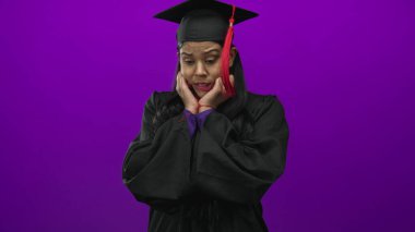 Mezuniyet kepi ve cüppeli, elleri mor stüdyoda yanaklı genç Latin kadın endişeli ifadeyi yakalıyor;.