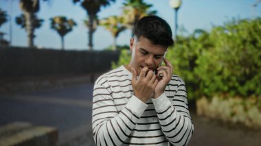 Genç adam telefon görüşmesi yaparken çizgili gömlek giymiş, palmiye ağaçlarının altında akıllı telefon kullanarak şehrin dışında dikiliyor..