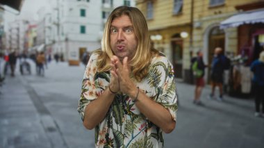 Tropikal tişörtlü sarışın adam kalabalık bir güneş ışığı sokağında kollarını kavuşturuyor ve titriyor..