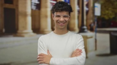 Kolları çaprazlanmış bir adam gözlük takıyor ve sokakta sütunlar inşa ederek göz kırparken gülümsüyor..