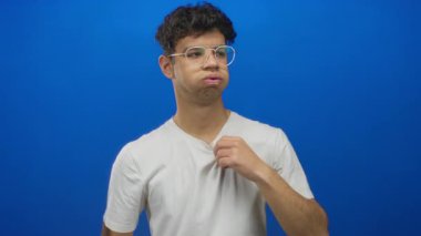 Temiz gözlüklü İspanyol genç adam mavi stüdyoda çıplak elle beyaz gömlek çekiyor..