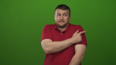 Yeşil bir stüdyoda eli kolu çapraz kırmızı bir polo giyen şişman bir adam, tereddütlüdür..