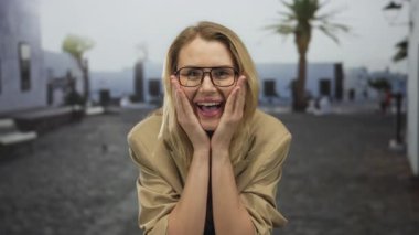 Güneşli bir sokak arkaplanında, yüzü açık gülümseyen bir kadın, sarı saçlı ve gözlüklü, sıradan bir ortamda heyecan ve neşe gösteren bir kadın.