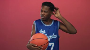 Mavi basketbol formalı genç adam basketbolu sıkıca tutuyor ve kırmızı stüdyoda kulaklarına dokunuyor..