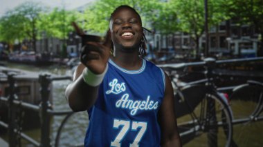 Adam gülümsüyor ve mavi basketbol forması içinde kameraya doğru işaret ediyor. Kanal korkulukları, Amsterdam şehri sahnesi, neşe yoldaşlığı..