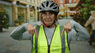Başlık ve güvenlik yeleği giyen yaşlı İspanyol kadın şehir sokaklarını göstererek şehir güvenliğini ve farkındalığı iletiyor..