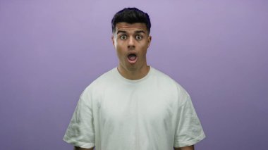 Genç adam, canlı bir mor arkaplan üzerine izole edilmiş, sıradan beyaz bir gömlek giyen, yüzün yanında elleri olan bir dizi duygu sergileyen, şaşırdığını ifade ediyor..