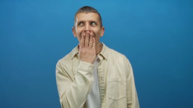 Şaşırmış görünen bir adam, mavi izole edilmiş arka plan karşısında elleri ve ağzını kapatır. Günlük gömlekler giyer, duygularını ve ifadesini ifade eder.