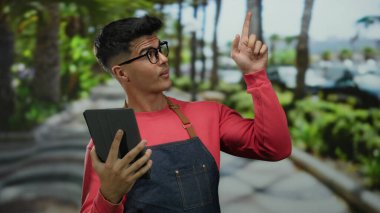 Gözlüklü genç adam dışarıda, yemyeşil bir park ortamında tablet kullanıyor, günlük kırmızı kıyafetler ve önlük giyiyor, odaklanmış ve dokunmatik ekran cihazıyla meşgul görünüyor..