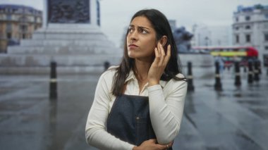 Woman holding hand to ear on wet street beside stone fountain and bollards, wearing denim apron, grimacing in pain; confusion.