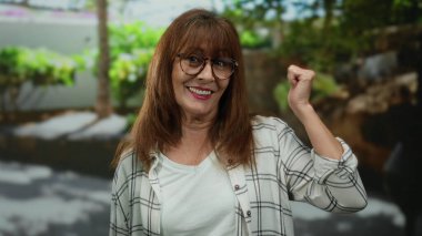 Kıdemli İspanyol kadın şehir caddesinde gülümsüyor açık havada olumlu bir jest yapıyor doğal yeşillik ile canlı şehir ortamında pozitifliği ima ediyor.