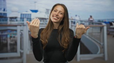 Kadın iki eliyle ve avuç içi açık bir şekilde yolcu gemisinin güvertesine çağırıyor, kameraya gülümsüyor; neşeyi davet ediyor.