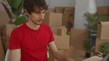 Kırmızı tişörtlü genç adam, karton kutular ve evrak kağıtlarıyla çevrili binadaki ahşap masada dizüstü bilgisayarda yazıyor..