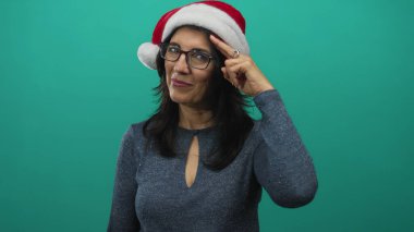 Woman wearing santa hat and glasses smiles and points her finger in green studio; holiday cheer.