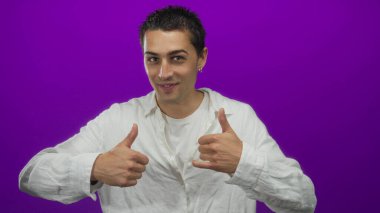 İzole edilmiş mor bir arka plan duvarı üzerinde neşeli bir ifadeyle başparmak kaldıran genç İspanyol..