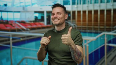 Genç bir adam canlı bir açık hava otel havuzunda başparmağını gösteriyor. Davet edilen su ve şezlongların arka planına güvenle gülümsüyor..