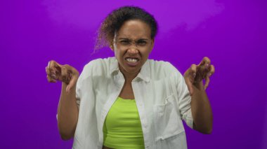 Beyaz bir gömlek ve neon yeşil bir üst giyen, canlı mor arka planı olan bir stüdyoda yüzünü buruşturan ve pençeleyen bir kadın..