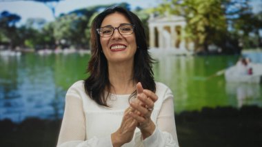 Orta yaşlı İspanyol kadın, güneş ışığı altında yeşil parkta gülümseyip el çırpıyor..