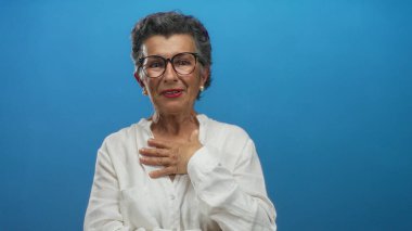 Beyaz bir bluz giyerken gri saçlı ve gözlüklü yaşlı bir kadın etkileyici pozlarında kendine güveni ve bilgeliği yayıyor..