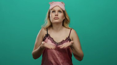 Pembe gecelikli bir kadın, uyku maskesi takmış, sakin bir ortamda yeşil bir zemine yaslanıyor ve nefes alıyor. Rahatlama ve farkındalık sergiliyor..