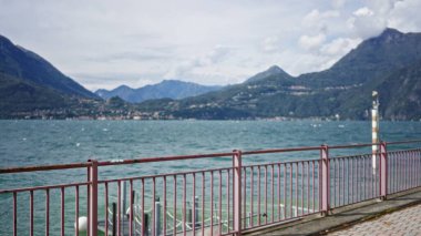 Paslı korkuluklar ve uzak dağlarla odaksız göl gezintisi, bulanık ön plan dış mekan; arkaplan telif alanı sakinliği.