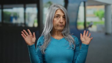 Son sınıf öğrencisi, gri saçlı, kapalı bir otel ortamında elini kaldıran mavi kazaklı, sıradan bir atmosferde derin düşüncelere dalmış bir yüz ifadesi sergiliyor..