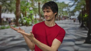 Kırmızı gömlekli bir adam, iki eliyle, palmiye ağaçlarıyla kaplı, güneşli bir şehir sokağını işaret ediyor..