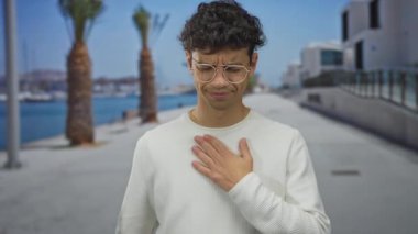 Deniz kenarındaki güvertede mavi denizin yanında palmiye ağaçları olan beyaz süveter giyen bir adam. Acı..