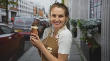 Önlükle gülümseyen bir kadın kalabalık bir şehir caddesinde dondurma külahı tutuyor, açık şehir ortamında neşe ve sıcaklık saçıyor..