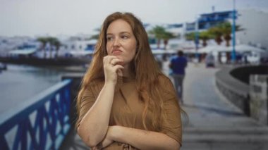 Deniz kenarında, sıcak güneş ışığı altında elini çenesine koyan beyaz bir kadın mesafeye bakıyor..