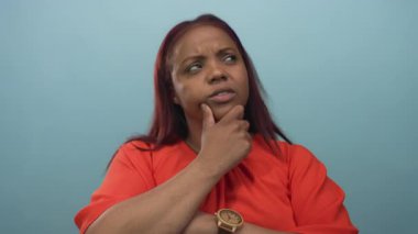 Eli çenesinde olan bir kadın stüdyoda soluk mavi bir arkaplan karşısında düşünceli bir poz veriyor; düşünceli bir yansıma.