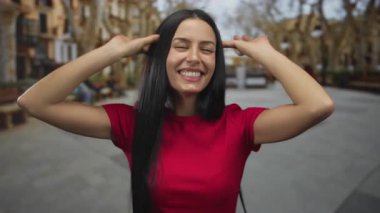 Kırmızı tişörtlü genç İspanyol kadın şehir sokaklarında neşeli yüzler sergiliyor. Arka planda şehir manzarası gösteren yayalar ve binalar var..