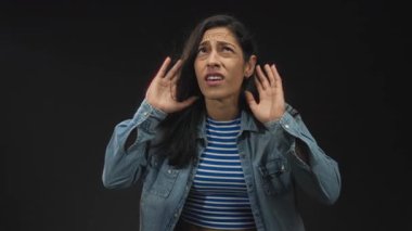 Stüdyoda elleri kulaklarında, siyah arka planda, çizgili bluz ve kot ceket giyen, duyusal aşırı yüklenme sorunu olan bir kadın..
