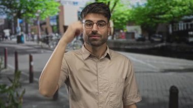 Bej gömlekli ve yuvarlak gözlüklü genç İspanyol adam sokakta kamerayı işaret ediyor..