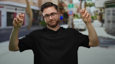Sakallı genç bir adam, şehir merkezinde gün ışığında dışarıda dikilirken gözlük ve günlük giysiler gösteren bir sokakta barış işareti yapıyor..