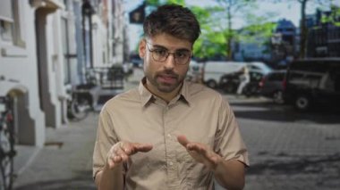 Bej gömlekli ve kahverengi gözlüklü adam şehir binalarının ortasında sakin bir jestle el ele tutuşuyor..