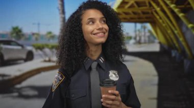 Dışarıda gülümseyen genç bayan polis memuru üniformalı bir şekilde sokakta kahve tutuyor.