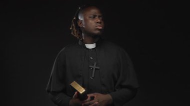 Siyah stüdyoda elleri göğsüne yakın yaldızlı İncil 'i tutan genç Afro-Amerikan rahip..